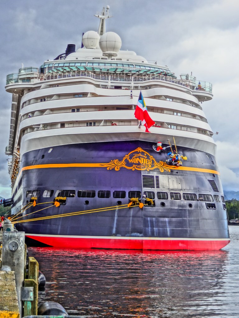 A large cruise ship docked at a port, featuring a red, black, and white exterior with a prominent flag mounted on the side.